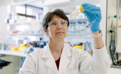 A woman stands in a laboratory wearing a white coat and glasses and holds up a glass vial